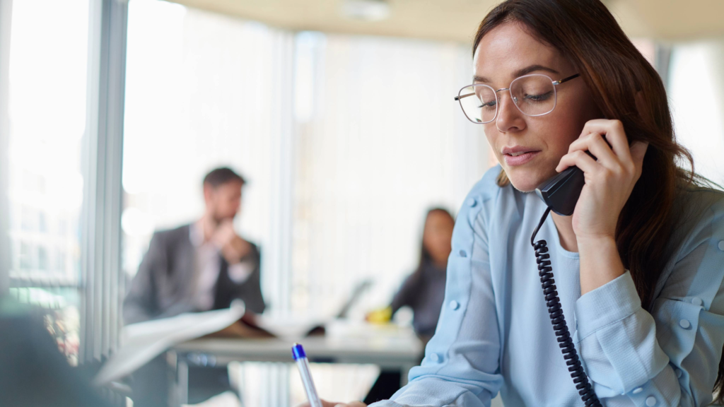 A woman in a blue blouse is on the phone, taking notes in an office setting. The image emphasizes the importance of getting new sales reps comfortable quickly with their tasks, while ensuring they have expert backup for handling complex technical details.