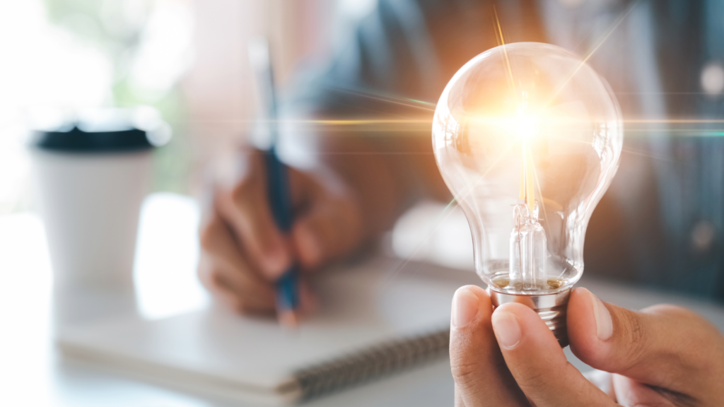 A bright lightbulb is being held by someone writing on a notepad, signifying a key insight being discovered as part of the customer interview.