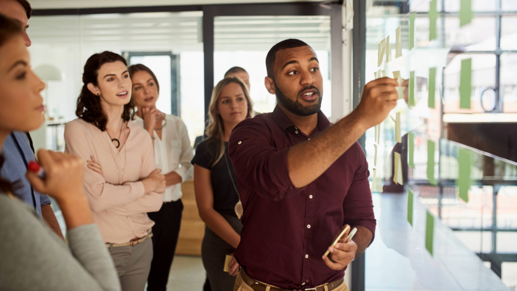 Business team collaborating as a man leads a strategy session, pointing to sticky notes on a glass wall
