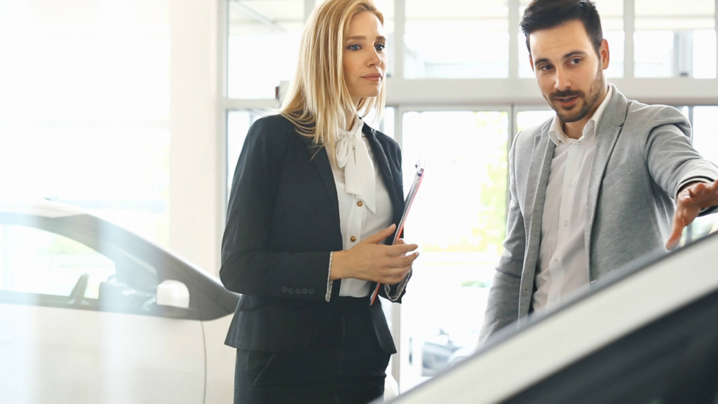 Salesperson explaining details of a car to a businesswoman holding a clipboard inside a bright showroom.